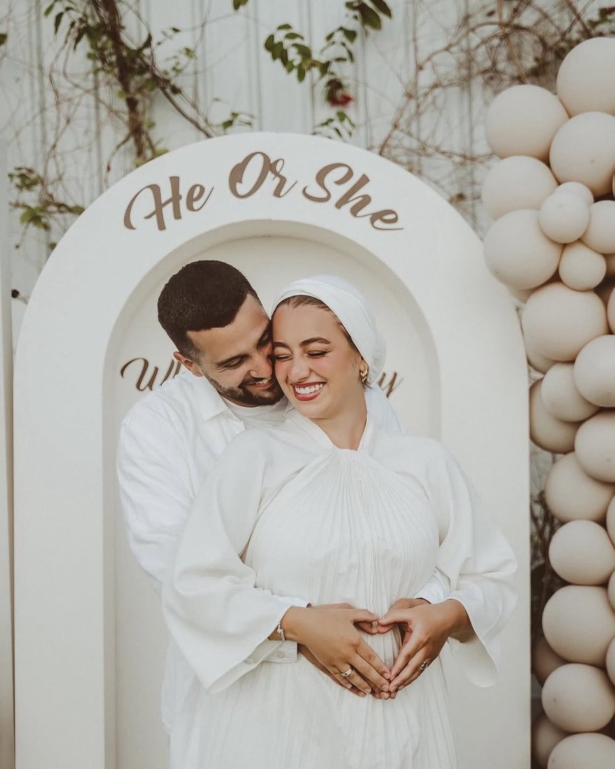 Couple at a white “He Or She” gender reveal arch with a neutral balloon installation by DT Events